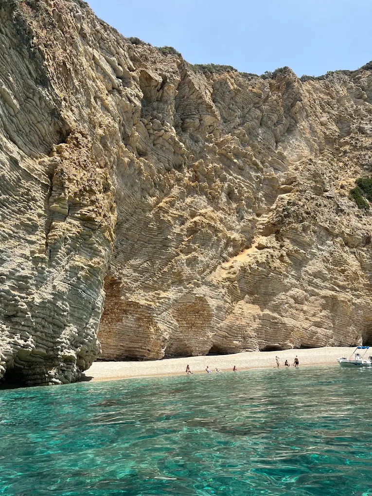 A tall limestone cliff on the Paleokastritsa coast dropping to a narrow pebble beach, seen from the water.